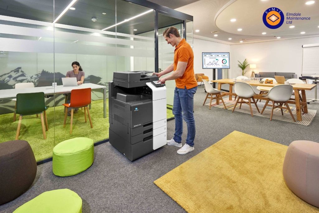 How to Choose the Best Managed Print Services Company | Elmdale Maintenance Ltd A modern office workspace featuring a Konica Minolta multifunction printer in the foreground. An employee wearing an orange polo shirt operates the printer while a colleague works on a laptop inside a glass meeting room. The open-plan office includes meeting tables, contemporary seating, a wall-mounted business analytics screen, and Elmdale Maintenance branding, illustrating a professional managed print environment.